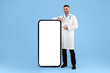 © Prostock-studio - Male physician in white uniform pointing finger at huge cellphone with blank screen on blue studio background, mockup