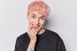 Horizontal shot of cheerful guy with dyed pink hair bites finger has happy expression wears casual black t shirt looks curiously isolated over white background. Positive human emotions concept