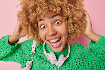 Wall Mural - Headshot of cheerful curly haired young woman looks surprisingly at camera smiles broadly wears green knitted jumper and stereo headphones around neck for listening music isolated over pink background