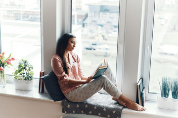 Sticker - Relaxed young woman in cozy pajamas reading a book while sitting on the window sill at home