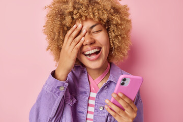 Wall Mural - Cheerful curly haired woman makes face palm smiles broadly shows white teeth holdds smartphone reads funny content dressed in fashionable purple jacket isolated over pink background. Positive emotions
