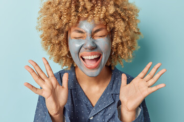 Wall Mural - Emotional curly haired woman applies facial mud clay mask keeps palms raised up exclaims loudly dressed in pajama undegoe beauty procedures at home after awakening isolated over blue background