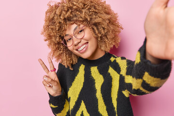 beautiful curly haired woman smiles gladfully shows peace sign feels carefree keeps arm outstreched 
