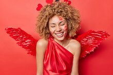 Curly Haired European Woman Smiles Positively Keeps Eyes Closed Has Traces Of Kiss On Face Wears Dress And Wings Behind Back Keeps Eyes Closed Isolated Over Red Background. Angel Female Model