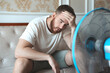 Young bearded man using electric fan at home, sitting on couch cooling off during hot weather, suffering from heat, high temperature