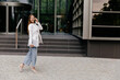 © PhotoBook - Full-length photo of stylish good-looking woman in white jacket and jeans wearing heels walking in city with phone and holding laptop