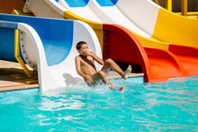 A Little Boy Slides Down A Water Slide And Has Fun. The Boy Swims In The Pool After Going Down The Water Slide In Summer.