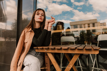 Caucasian young pretty stylish woman sitting table near street cafe restaurant chill dressed brown trench coat smiling happy poses outside in city, spring autumn season. Cute brunette hair lady

