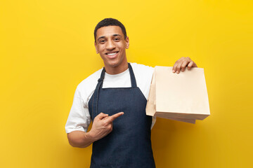 Poster - young African American barista guy in uniform holds packages of food on yellow background, delivery service