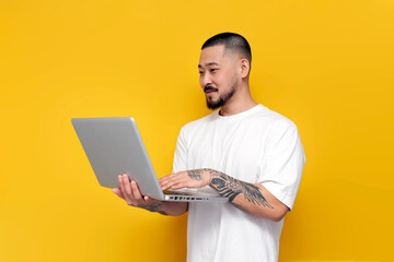 Wall Mural - adult tattooed asian man in white t-shirt uses laptop on yellow isolated background, unshaven korean guy