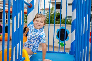 Little preschool girl sliding on a children slide in outdoor swimming pool in hotel resort. Child learning to swim in outdoor pool, splashing with water, laughing and having fun. Family vacations.