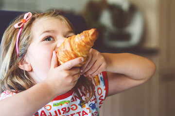 Wall Mural - Smiling child at breakfast. Food and happy kids. The girl is eating a croissant. Cute preschool girl having healthy meal.