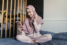 Smiling Young Woman Wearing Hijab Text Messaging Using Smart Phone In Bedroom