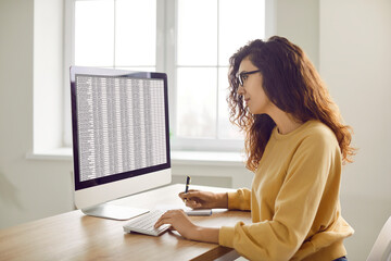 Female accountant working with digital business spreadsheets on office computer. Young woman in casual yellow jumper sitting at desk, looking at screen, and using modern accounting software