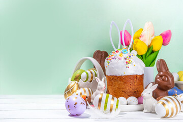 Wall Mural - Tasty Glazed Easter cake with sugar decor, colorful eggs basket, ceramic rabbits and spring flower on white kitchen table, copy space. Happy Easter Holidays greeting card background copy space