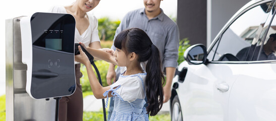 Progressive young parent teach daughter how to recharge or refuel EV car at home charging station. Green and clean energy from electric vehicle for healthy environment. Eco power from renewable source