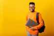 © Vadim Pastuh - Happy excited foreign male student with backpack standing isolated on yellow and holding laptop, arab guy in glasses ready to studying, back to school concept