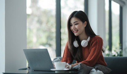 Wall Mural - asian young woman listening music with headphone and streaming music from laptop on sofa relaxing at home