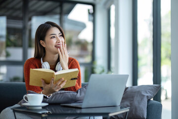 Wall Mural - asian young woman listening music with headphone and streaming music from laptop on sofa relaxing at home