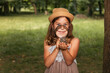 © _KUBE_ - Children's education and curiosity. Closeup portrait of caucasian smiling little girl in a straw hat looks at pine cone through a magnifier. The concept of scouting and happy childhood