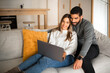 © Prostock-studio - Smiling international millennial couple sit on sofa, hugging, looking at laptop, have video call in living room