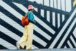 © okrasiuk - Back view stylish woman in bright clothes and bucket hat wearing wireless headphones and listening to music while walking near city urban painted wall. Fashionable hipster lifestyle. Selective focus.