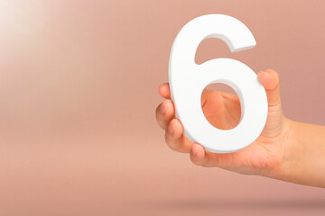 Fototapete - Number six in hand. A hand holds a white number 6 on a red background with copy space. Concept with number six. 6 percent, birthday 6, sixth grade, in sixth grade