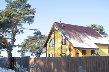 A yellow cozy house in the snow in winter in the village is surrounded by pine trees. Snow-covered roof, heating and ventilation pipes, trapezoid windows