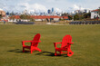 Two red chairs are sitting on green grass at The Presidio's Tunnel Top Park. In the background is the San Francisco cityscape. There is a blue sky with white clouds.