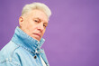 Serious aged man with white hair in blue denim jacket looking at camera while standing in isolation over vivid lavender background