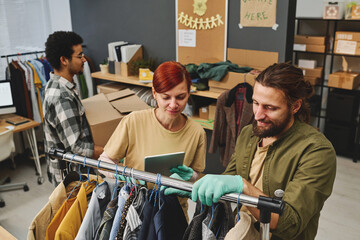 Young female volunteer with digital tablet making list of second hand clothes hanging on rack while bearded man helping her