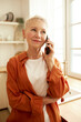 © Anatoliy Karlyuk - Vertical portrait of aged female in stylish orange shirt talking on phone to food delivery service, making order, standing against window at kitchen, looking aside with smiling face