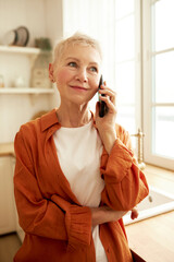 Wall Mural - Vertical portrait of aged female in stylish orange shirt talking on phone to food delivery service, making order, standing against window at kitchen, looking aside with smiling face