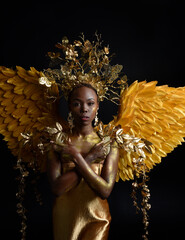 Wall Mural -  fantasy portrait of beautiful african woman model with afro, goddess silk robes, ornate crown & gold angel wings.  gestural Posing holding golden flowers., isolated on dark  studio background 