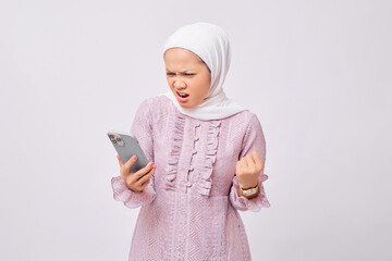 Angry beautiful young Asian Muslim woman wearing hijab and purple dress  screaming loudly into mobile phone isolated on white studio background