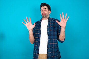 Wall Mural - Young man standing over blue studio background afraid and terrified with fear expression stop gesture with hands, shouting in shock. Panic concept.