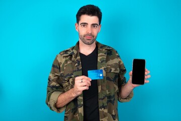 Photo of adorable Young man standing over blue studio background holding credit card and Smartphone. Reserved for online purchases