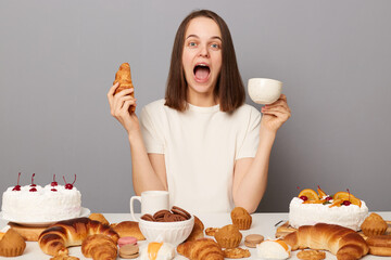 Wall Mural - Portrait of excited amazed woman with brown hair sitting at table isolated over gray background, breaking diet, drinking coffee or tea with yummy sugary croissant.