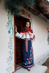 Wall Mural - Charming smiling woman in traditional ukrainian handkerchief, necklace and embroidered dress standing at background of decorated hut. Ukraine, style, folk, ethnic culture