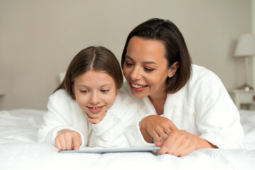 Wall Mural - Cheerful european millennial woman and little girl in bathrobes watch video on tablet, lie on soft bed