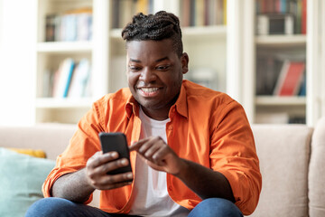 Wall Mural - Positive cool overweight black guy using smartphone at home