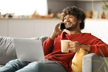 Wall Mural - Young Indian Man Talking On Mobile Phone And Using Laptop At Home