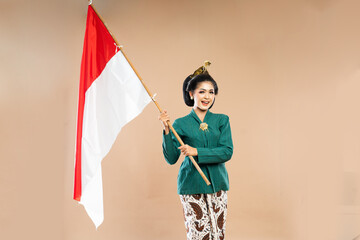 Wall Mural - asian woman in green kebaya standing with smile while holding the indonesian flag with her both hands on isolated background