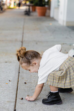 happy toddler girl in skirt picking up acorn from ground on street in Miami.