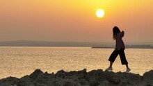 Aerial View Of A Happy Smiling Woman Jumping On Rocks On The Ocean Shore At Sunset. Freedom Smile Happiness Concept, Girl Fooling Around Like A Child. High Quality 4k Footage