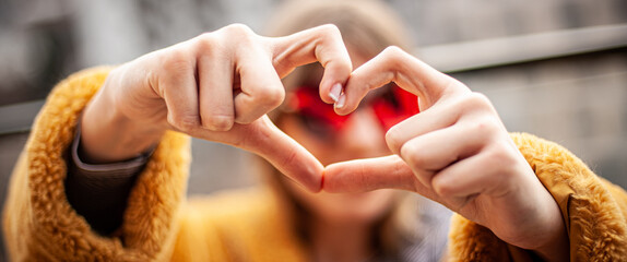 Capture Moments. Young girl in a yellow fur coat and red glasses making a heart shape with her hands and fingers, city in background.