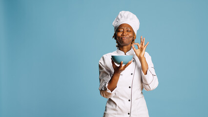 Wall Mural - Cheerful gourmet chef smelling food aroma from bowl on camera, enjoying professional meal cooked. Professional female cook savoring tasty food, flavor and scent in studio. Catering service.