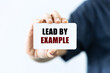 Lead by example text on blank business card being held by a woman's hand with blurred background. Business concept about leading by example.