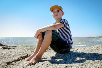 young preteen boy in striped shirt sitting on sandy beach with calm waves in the background