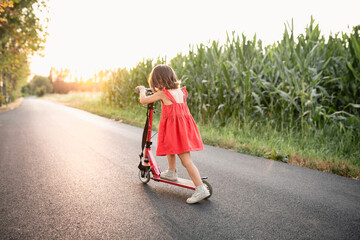 Wall Mural - girl in red dress riding red scooter on a road next to corn field